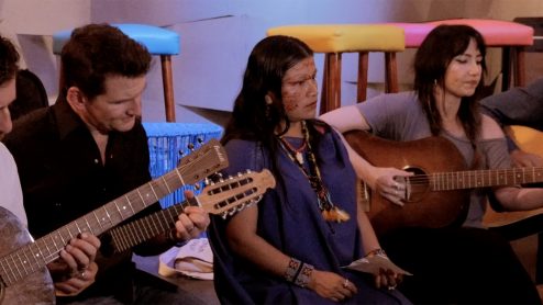 Musicians play guitars next to a tattooed person in Indigenous clothing