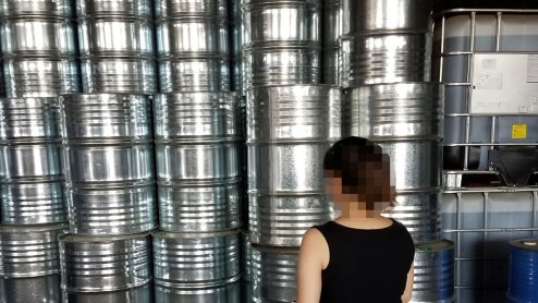 A person with face censored stands in front of a wall of silver chemical drums