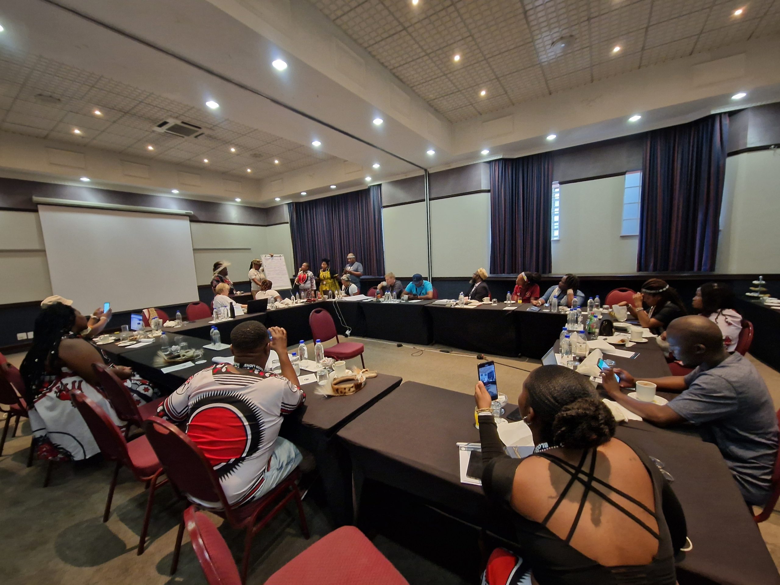 A wide view of the workshop room. People in chairs listen as a group near the front speak