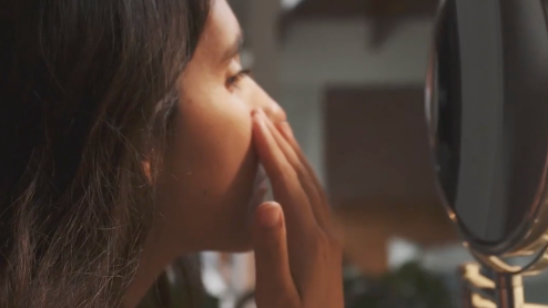 A woman with brown skin touches her face while looking in the mirror
