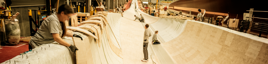 A group of workers stand in the middle of a wind turbine blade in the process of being built. The long wooden passageway is wide enough for multiple people to stand across and extends far into the distance