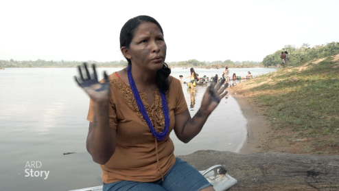 A person with painted face and hands talks to the camera while sitting next to a river